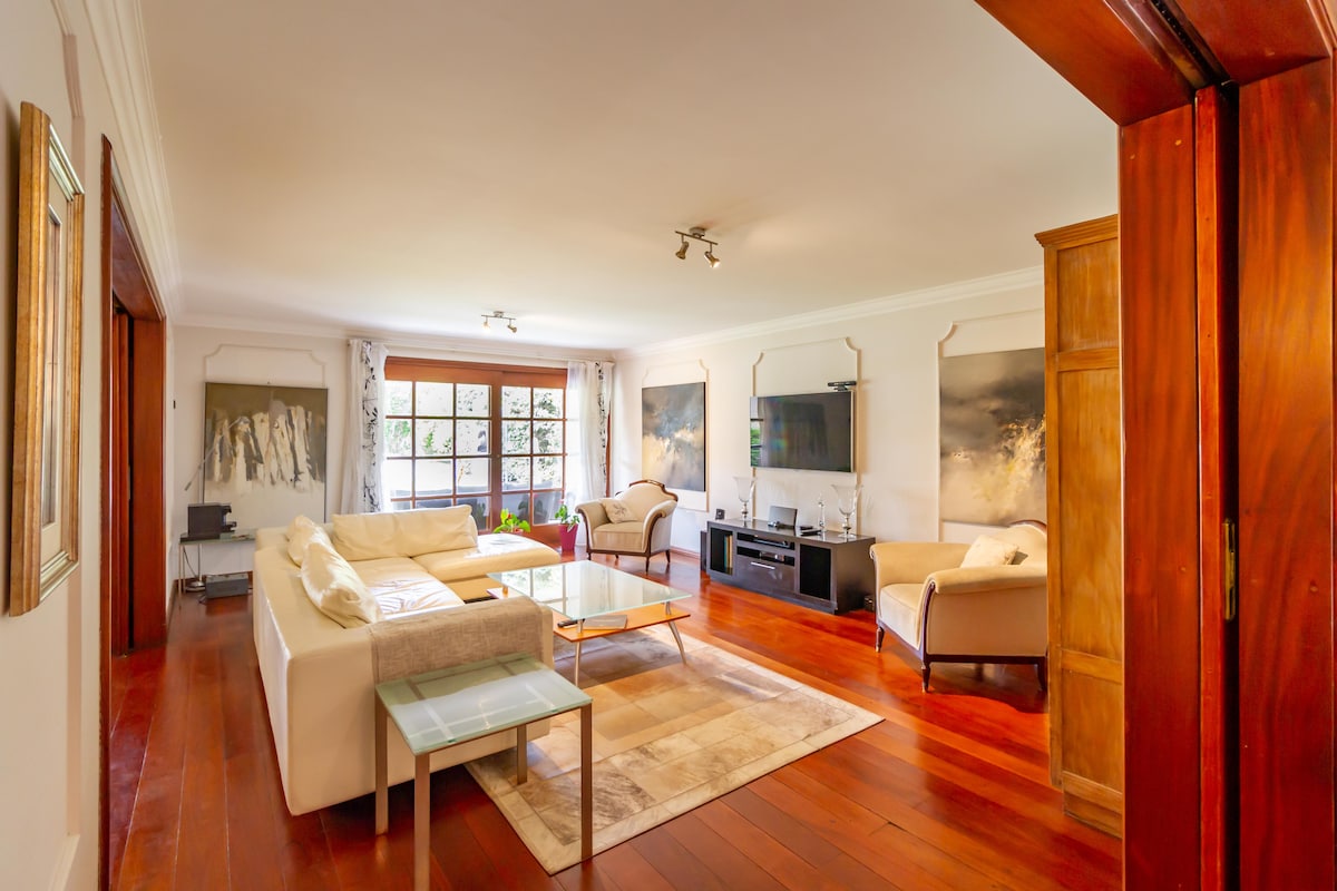 The spacious living room features hardwood floors and large windows allowing natural light to fill the space. Two comfortable armchairs and a plush sofa surround a glass coffee table. Two wall-mounted televisions are present, along with abstract art on the walls.