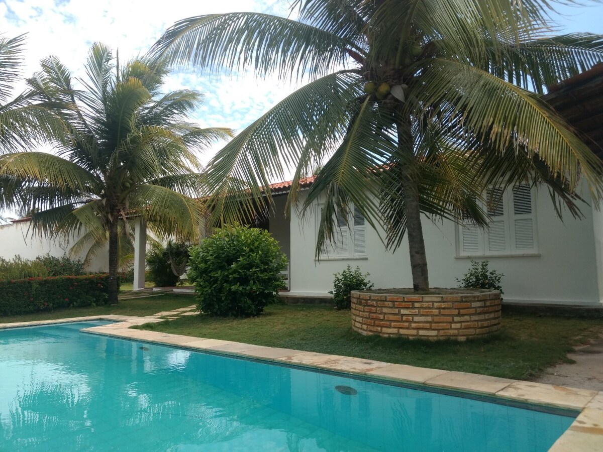 A tranquil view of a large swimming pool is bordered by lush greenery and palm trees, creating a serene outdoor space. A well-maintained garden complements the property, with a white-walled house visible in the background.