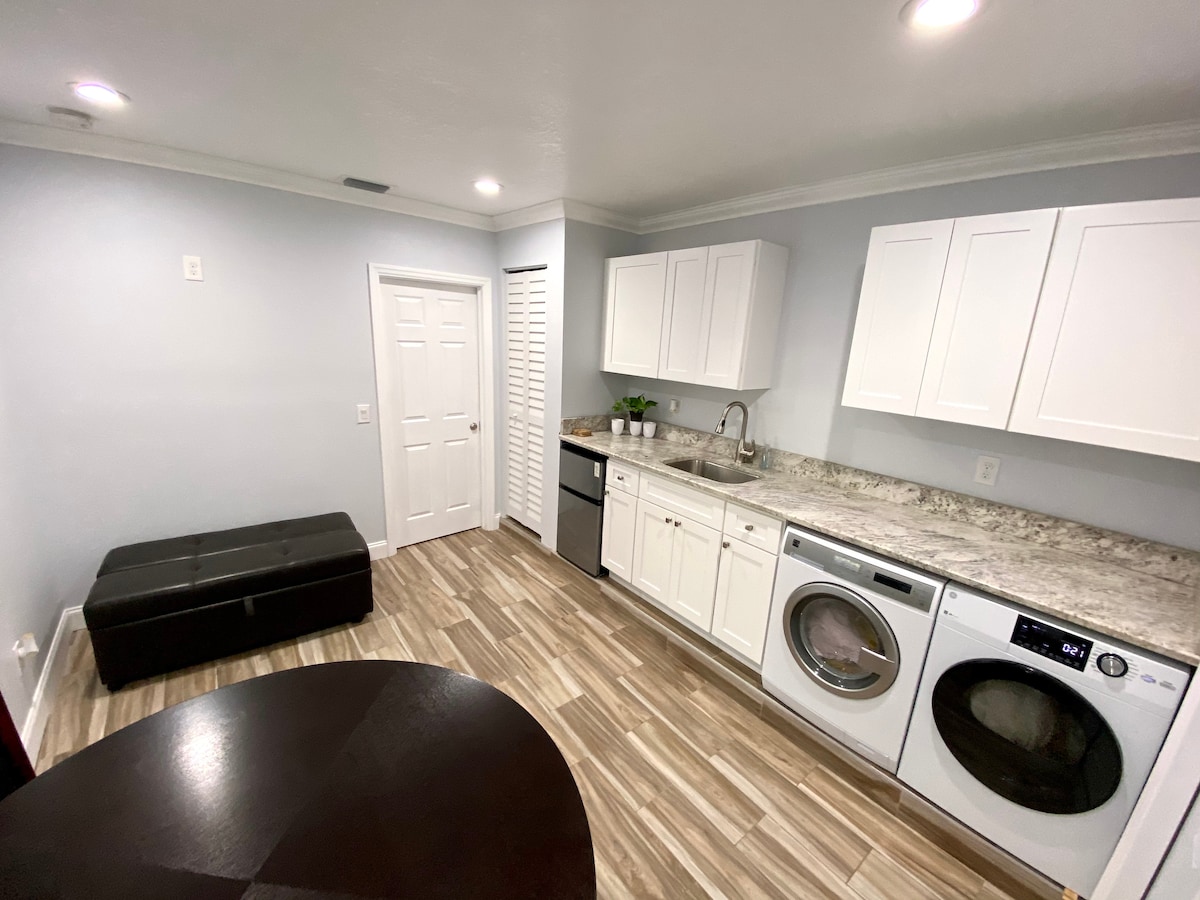 The kitchen area features white cabinetry and a granite countertop, accompanied by a mini-fridge, microwave, and a coffee maker. A washer and dryer are integrated into the cabinetry. A dark upholstered bench is positioned nearby, enhancing the functional space.