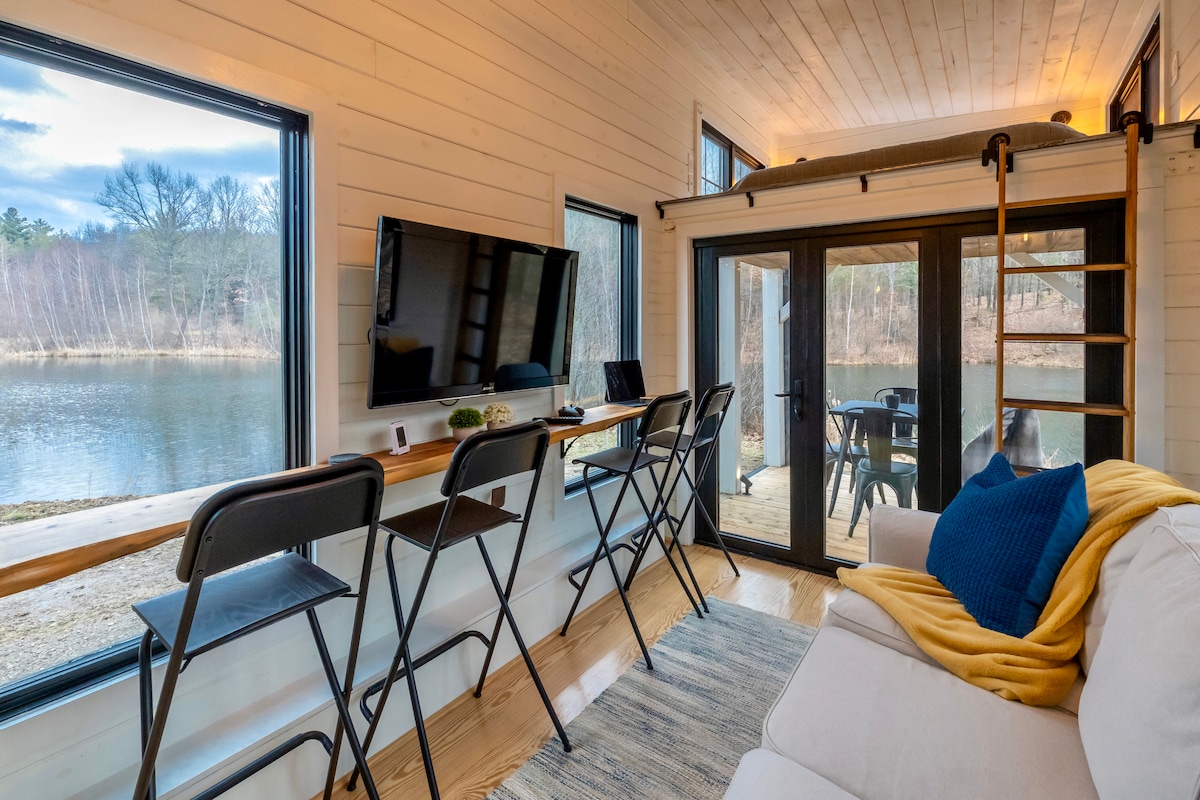 A bright, airy living area features large windows that provide views of the waterfront. A comfortable couch and a bar with four black folding chairs are visible, along with a mounted television. Natural light fills the space, enhancing the inviting design.