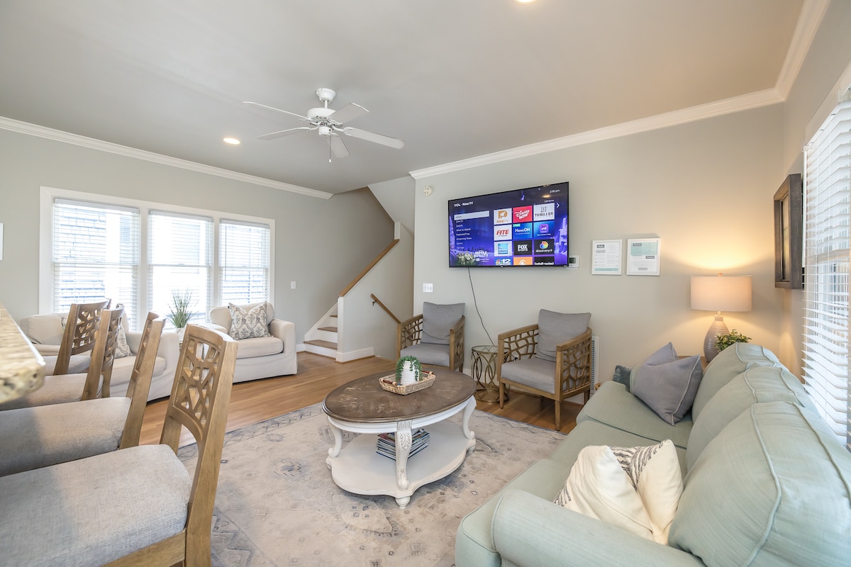 The living area features a combination of comfortable seating, including a soft blue couch and beige armchairs. A coffee table sits centrally, enhancing the inviting setup. A wall-mounted TV is visible, with stairs leading to an upper level in the background. Natural light enters through multiple windows.