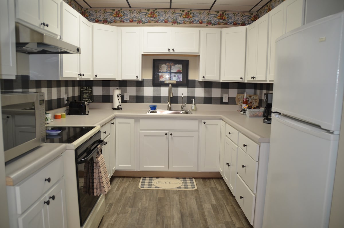 A fully equipped kitchen features modern appliances, including a stove, refrigerator, and microwave. White cabinetry and checkered patterns create a clean aesthetic. Countertops are organized, with cooking utensils readily available. A small rug lies near the sink, adding a touch of comfort to the space.