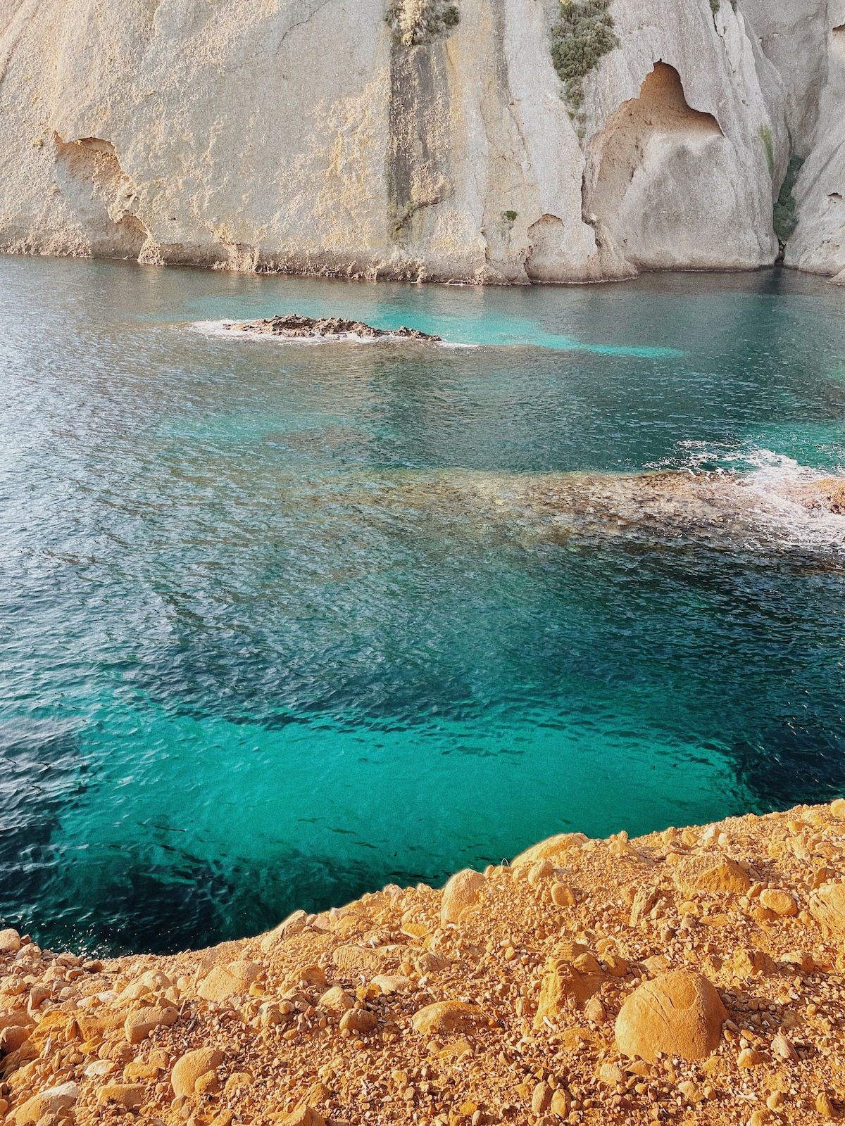 The image captures a serene coastal scene featuring a rocky shoreline with smooth, golden stones. Clear turquoise waters reflect the nearby cliffs, which have weathered textures and natural formations. The tranquil water is contrasted by the striking rock face, creating a harmonious seascape.