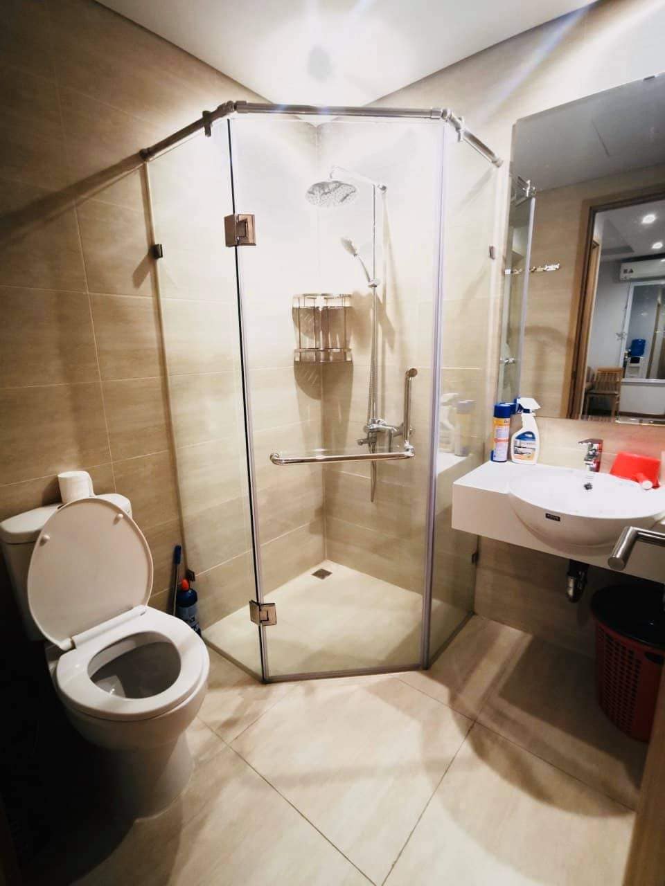 An enclosed shower area features clear glass walls, with a rainfall showerhead mounted above. A countertop sink is positioned next to it, complemented by a wall mirror. The floor is tiled in a light tone, creating a clean and modern appearance.