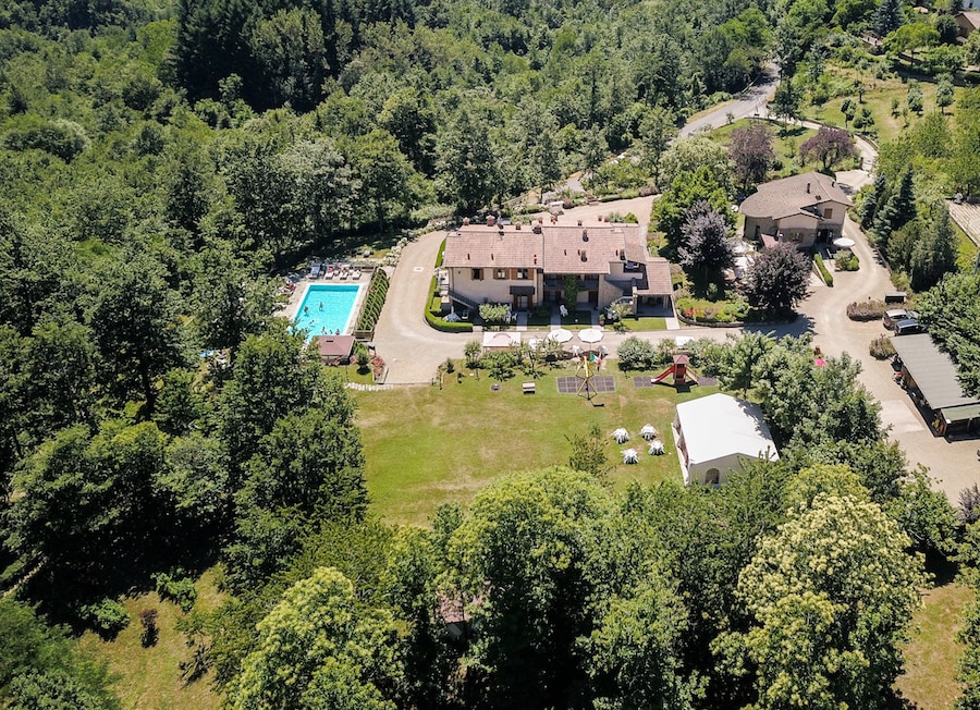 Aerial view of an agriturismo set amidst lush greenery, featuring a clear swimming pool surrounded by sun loungers. Several buildings are arranged along a winding path, with outdoor seating areas visible under shaded trees, creating a serene and spacious environment.