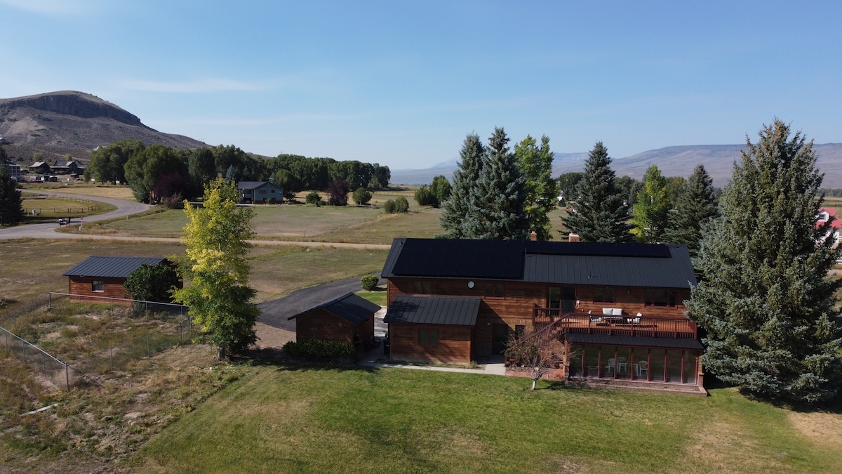 An aerial view captures a spacious wooden house with a dark roof, surrounded by a green lawn and tall trees. The structure sits in a rural landscape with mountains in the background and various homes in the distance, showcasing a peaceful environment.
