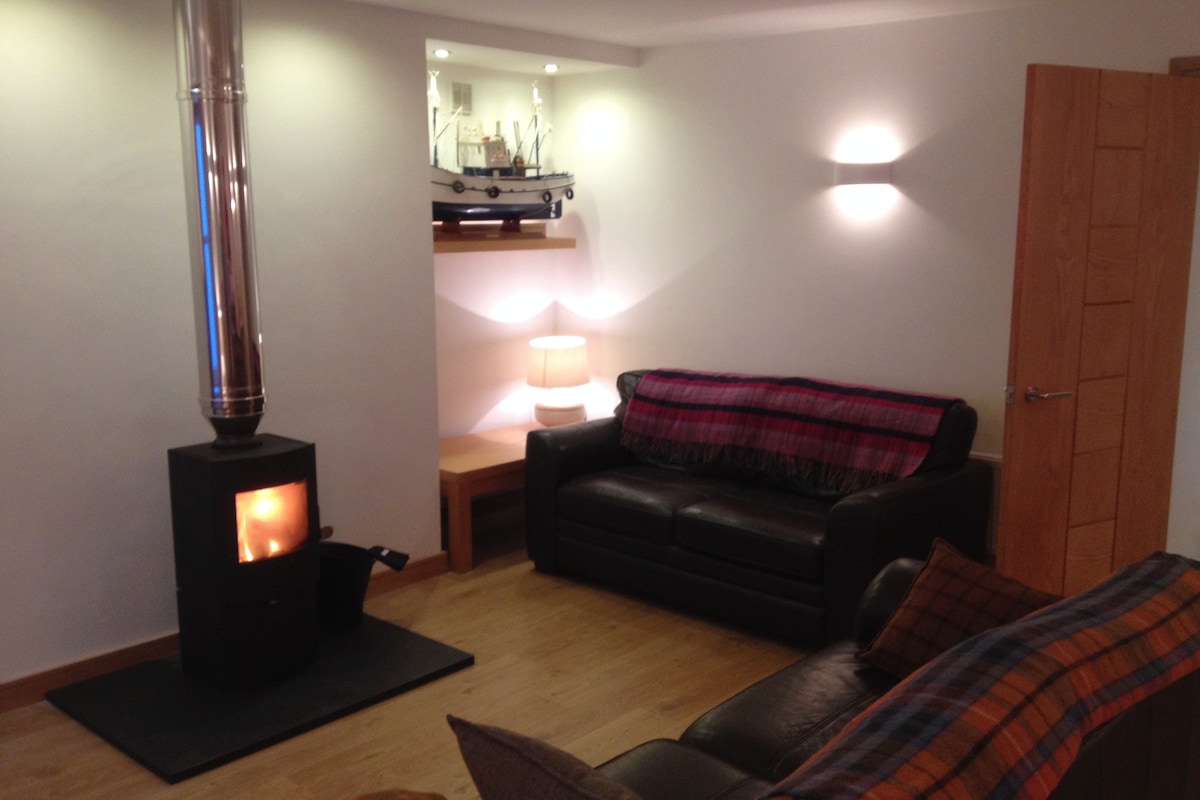 A warm living area features a wood burner set within a black metal surround, radiating a soft glow. A leather sofa is adorned with a colorful plaid throw, and a small shelf above displays a model boat and soft lighting, creating a cozy ambiance.