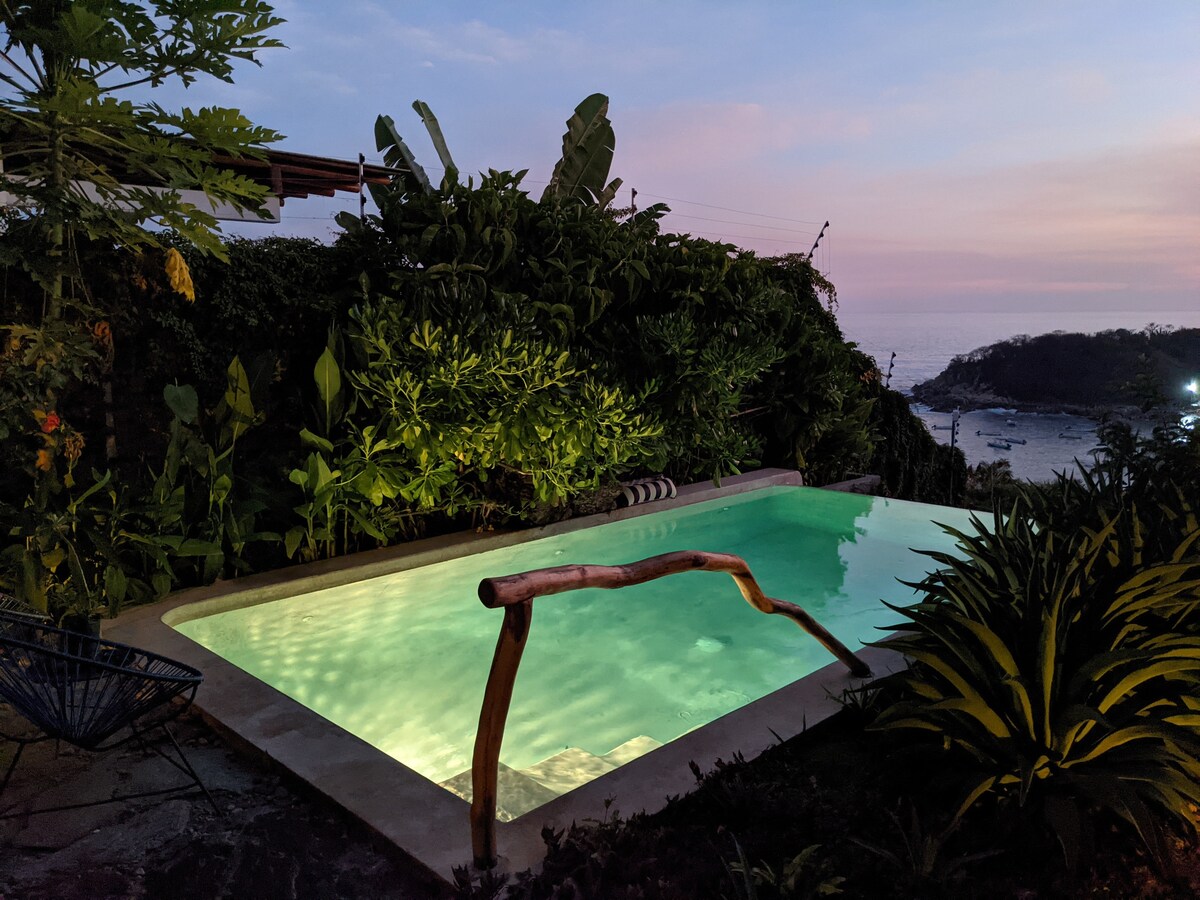 A refreshing salt-water pool is surrounded by lush greenery, with a wooden railing providing natural contrast. The pool's tranquil waters reflect soft evening light, creating a serene atmosphere. The view beyond includes distant ocean waves and gently sloping hills.