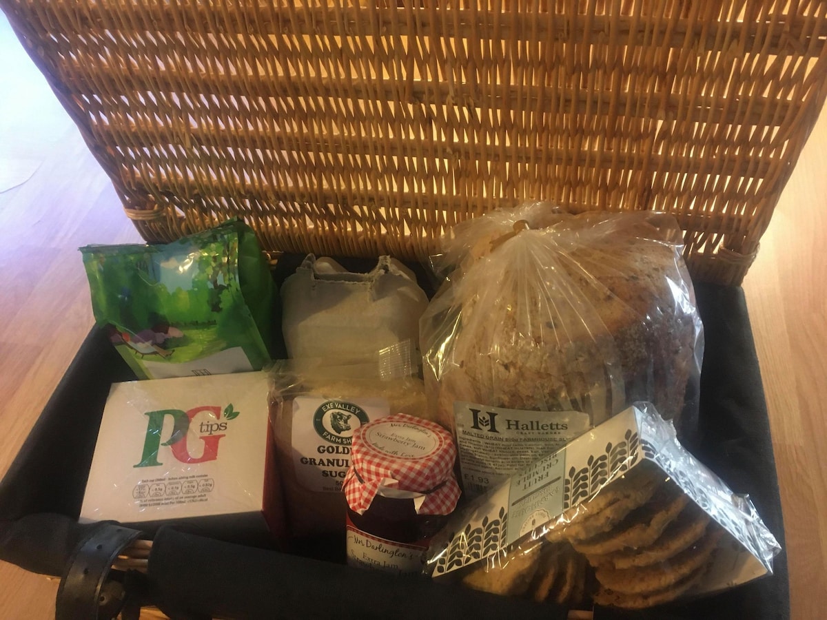 A woven basket filled with various food items is displayed. Inside, there are packets of tea, granulated sugar, a jar of strawberry jam, and a loaf of bread. Additionally, homemade cookies are presented in a clear package, offering a delightful welcome.