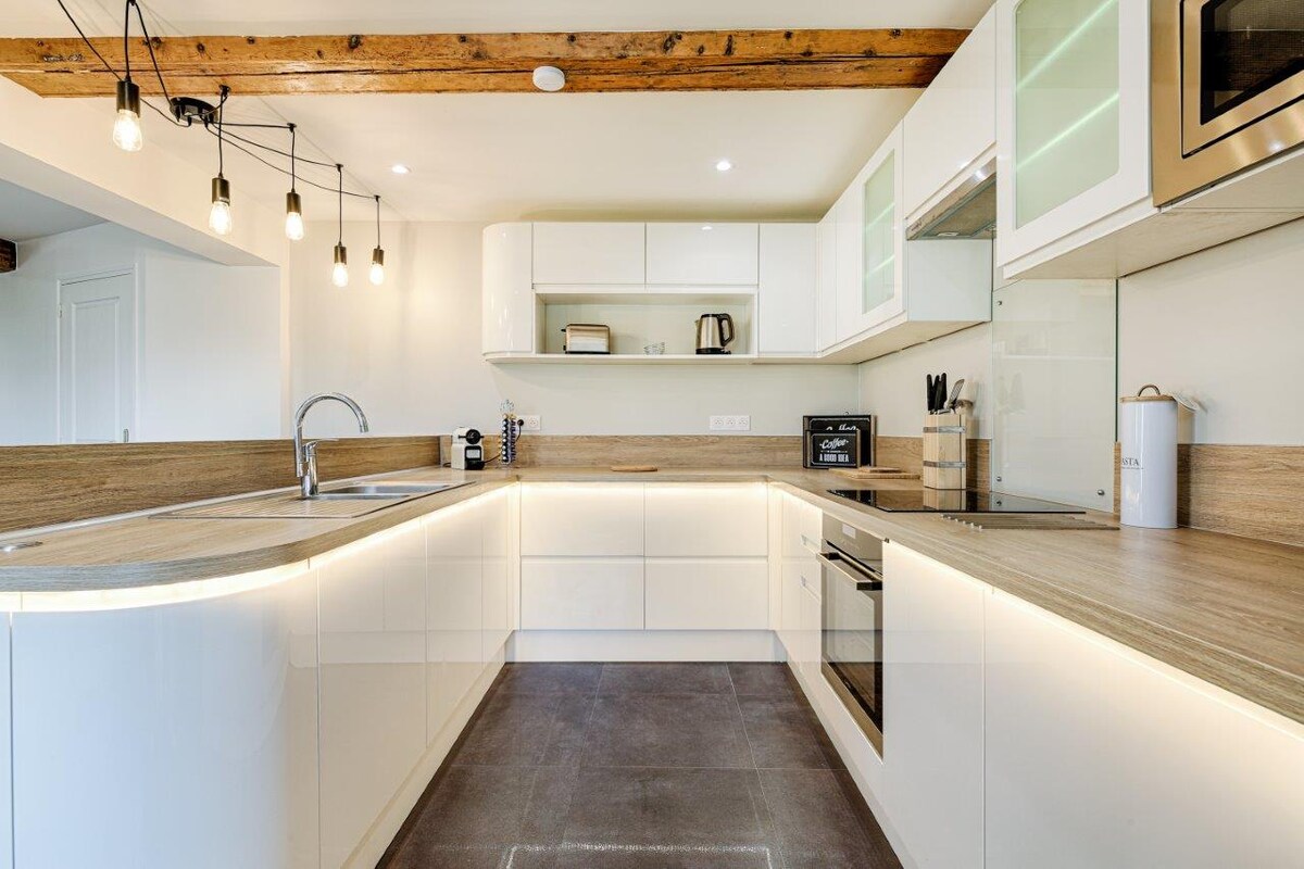 A modern kitchen equipped with sleek white cabinetry and wooden countertops features a unique curved layout. Soft under-cabinet lighting enhances the workspace. Stainless steel appliances, including an oven and kettle, complement the minimalist design, while exposed wooden beams add a touch of character.
