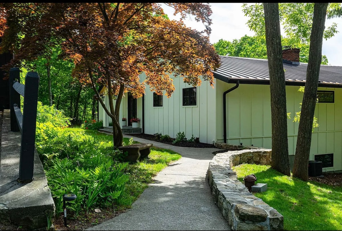 A winding stone pathway leads to a modern house, surrounded by lush greenery and vibrant foliage. The entrance is framed by a decorative maple tree and natural stone walls, creating a welcoming approach to the residence.