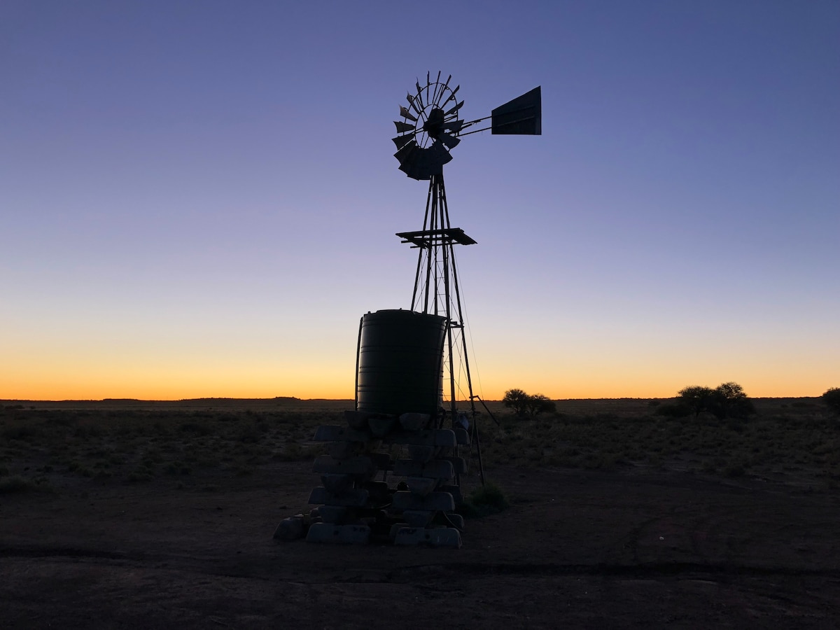 A silhouette of a windmill stands against a gradient sky at sunset, featuring hues of orange and blue. The structure is accompanied by a water tank supported by a stone base. The surrounding landscape is open, enhancing the sense of space and tranquility.