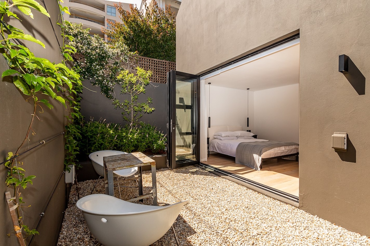 The image showcases a private outdoor space featuring a gravel surface and two relaxing bath-like chairs. A small wooden table complements the area, while large glass doors open to reveal a well-lit bedroom with a spacious bed and minimalist decor.