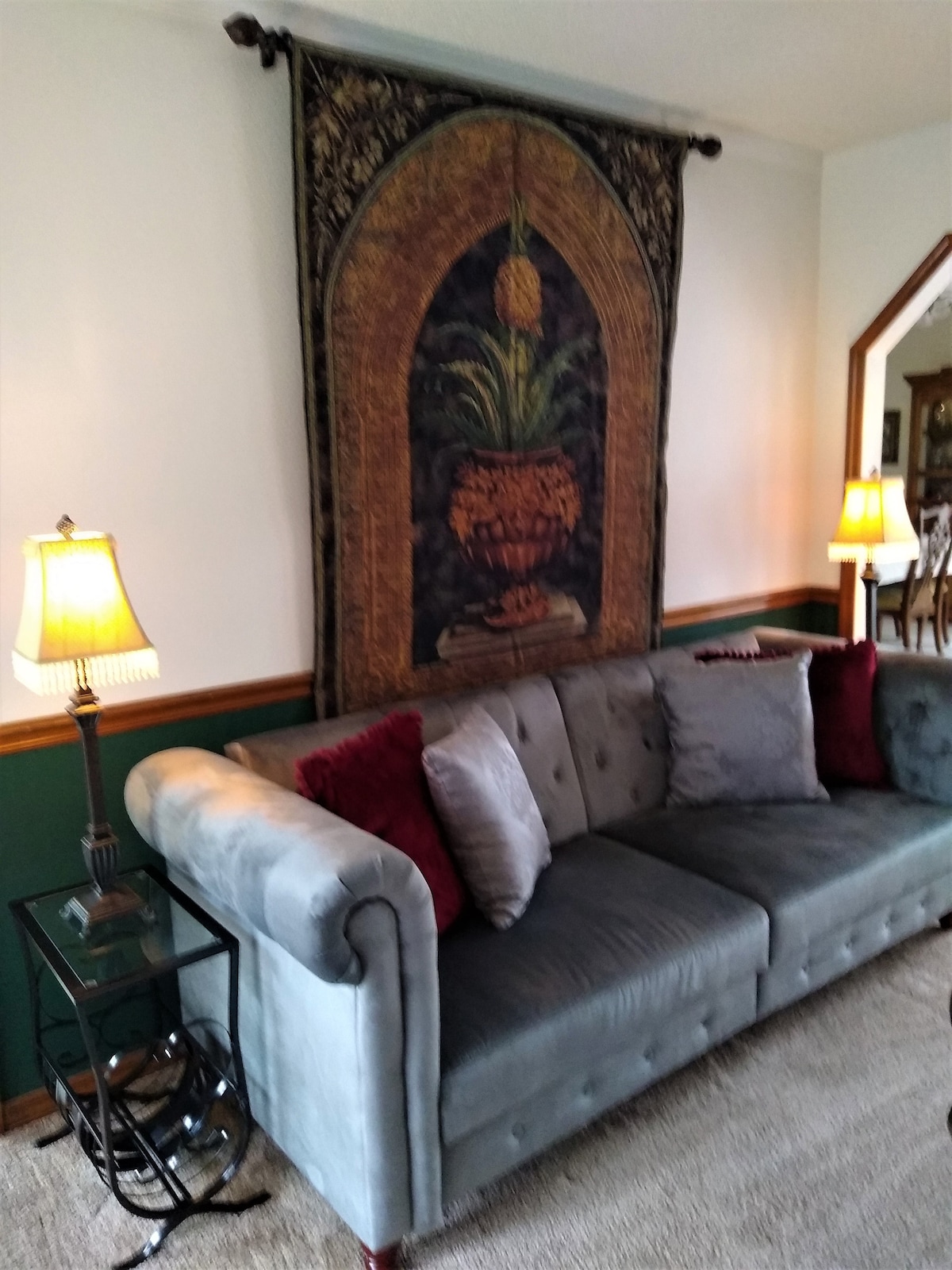 A tufted gray sofa is positioned against a pale wall, adorned with a decorative tapestry featuring a plant and urn. Two lamps provide soft lighting on either side of the couch, complemented by a side table holding a decorative item. Soft carpeting enhances the cozy atmosphere.