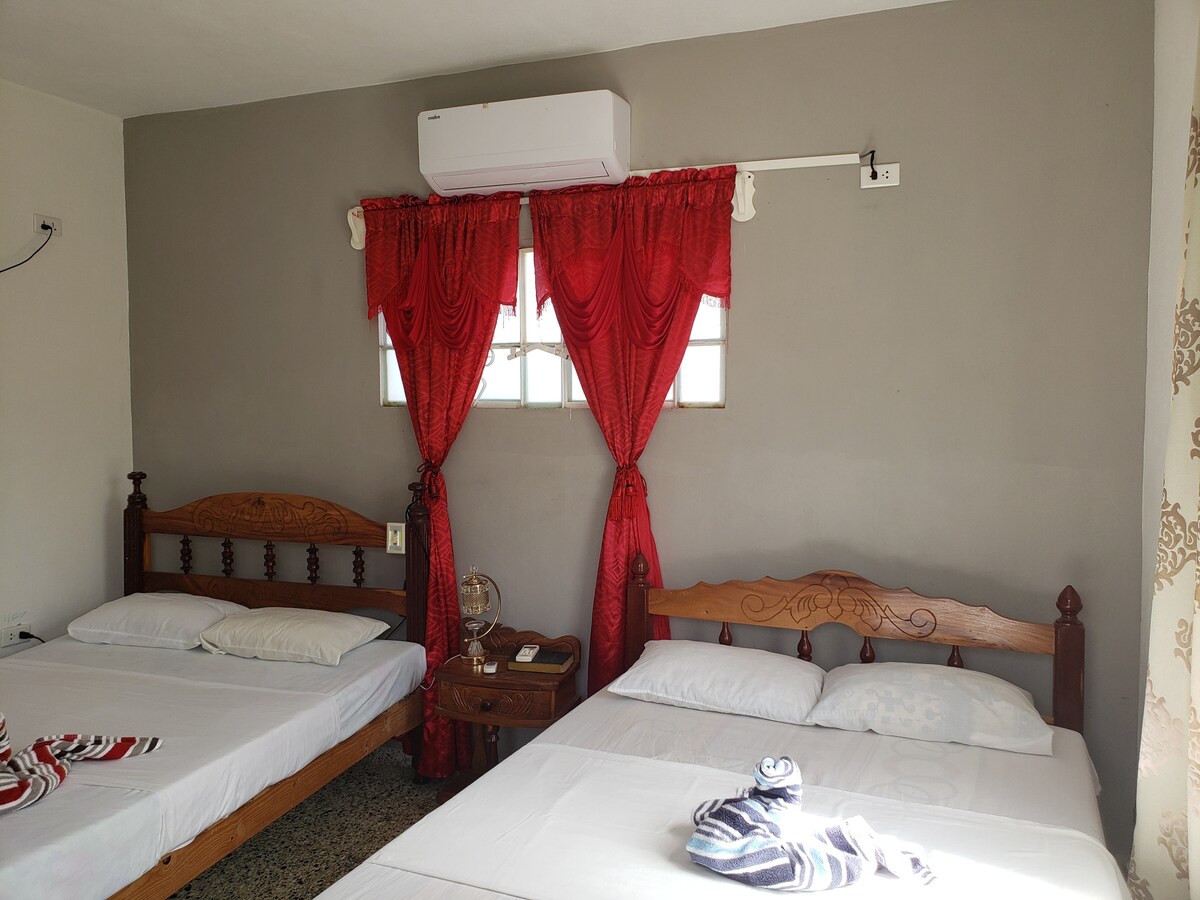 The room features two spacious beds with simple white linens. A decorative red curtain frames a window, allowing natural light to enter. A small wooden table beside the beds adds to the room's functionality, while an air conditioning unit is mounted on the wall for comfort.