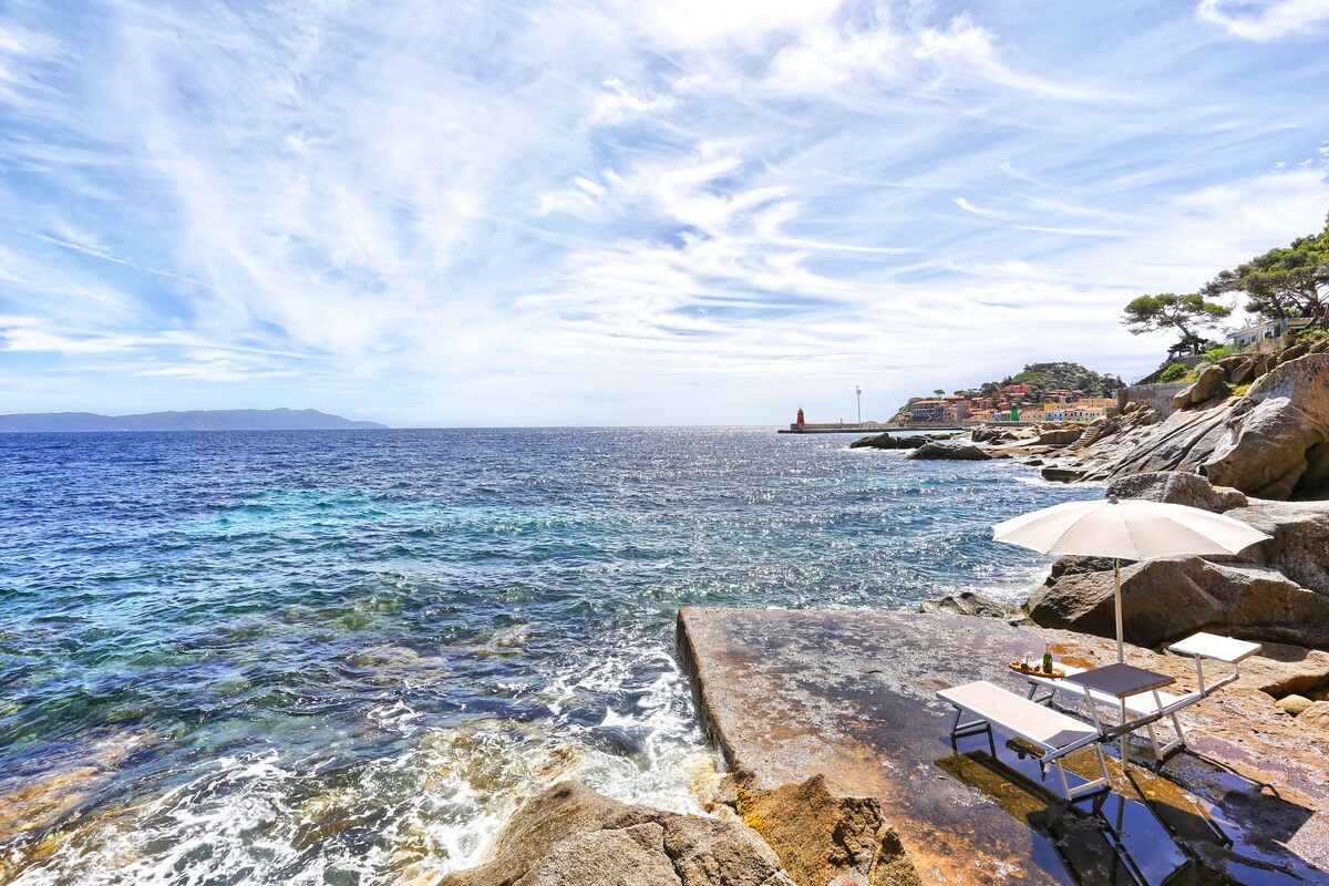 A rocky shoreline extends into clear blue waters, with a sunlit terrace featuring a lounge chair and a large umbrella. In the distance, coastal buildings and a lighthouse are visible against a backdrop of wispy clouds and blue sky.