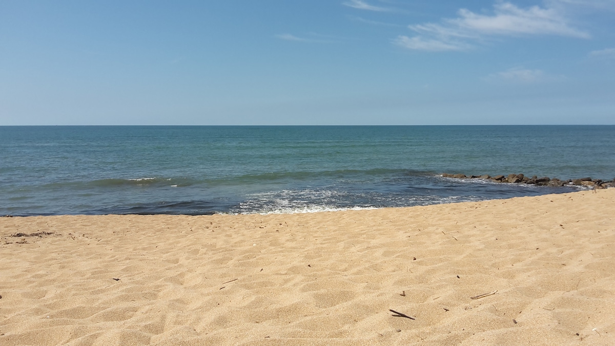 A serene beach scene features golden sand extending towards gentle waves lapping at the shoreline. The calm blue water meets a clear sky, creating a tranquil atmosphere. Rocky formations are visible at the water's edge, adding natural texture to the coastal landscape.