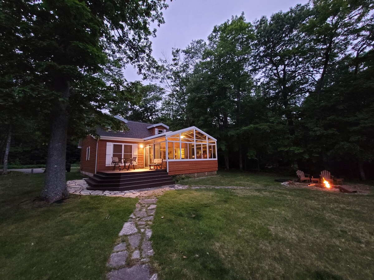 The exterior of the property is showcased with warm lighting emanating from the windows. A stone pathway leads to the seating area on the deck, while a fire pit sits surrounded by outdoor seating in the yard, all set against a backdrop of lush trees.