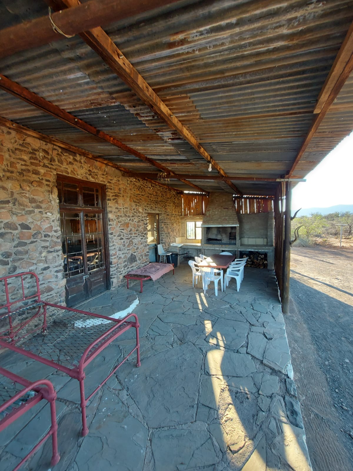 An outdoor seating area is presented with stone flooring, shaded by a rustic overhang. A simple wooden table and white chairs are arranged for dining, with a brick-built braai area visible in the background. Red metal benches add color to the setting.