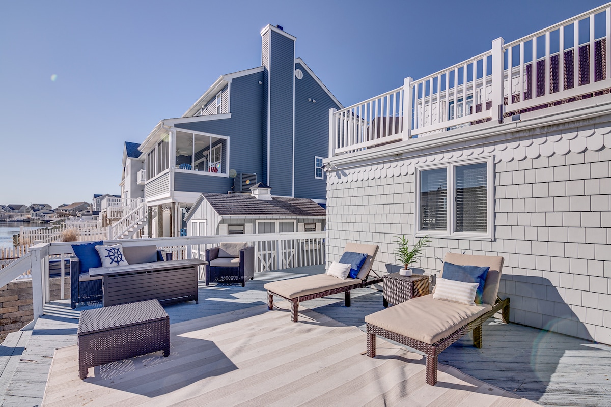An outdoor deck features comfortable loungers and a small table, creating a relaxing space for guests. The area is complemented by views of the waterfront and a nearby house. The deck is designed for leisure and enjoyment of the surrounding scenery.