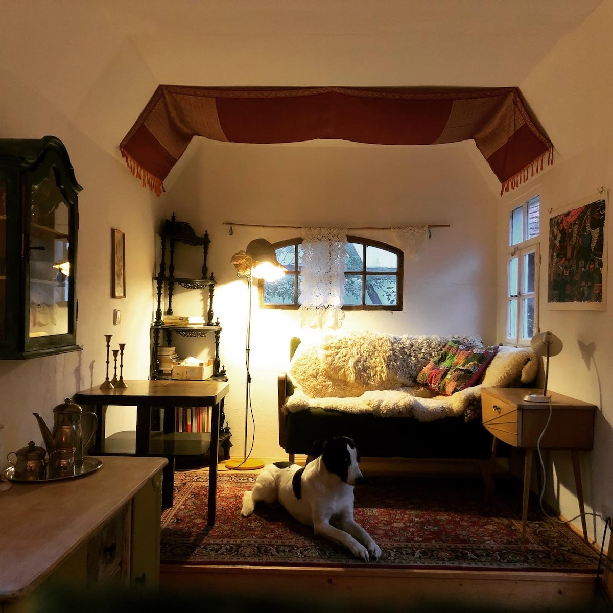 A cozy interior space features a charming seating area with a dark sofa adorned with decorative pillows and throws. An eclectic side table complements the setting, while a soft rug covers the wooden floor. A dog relaxes nearby, and warm light from a standing lamp illuminates the room.