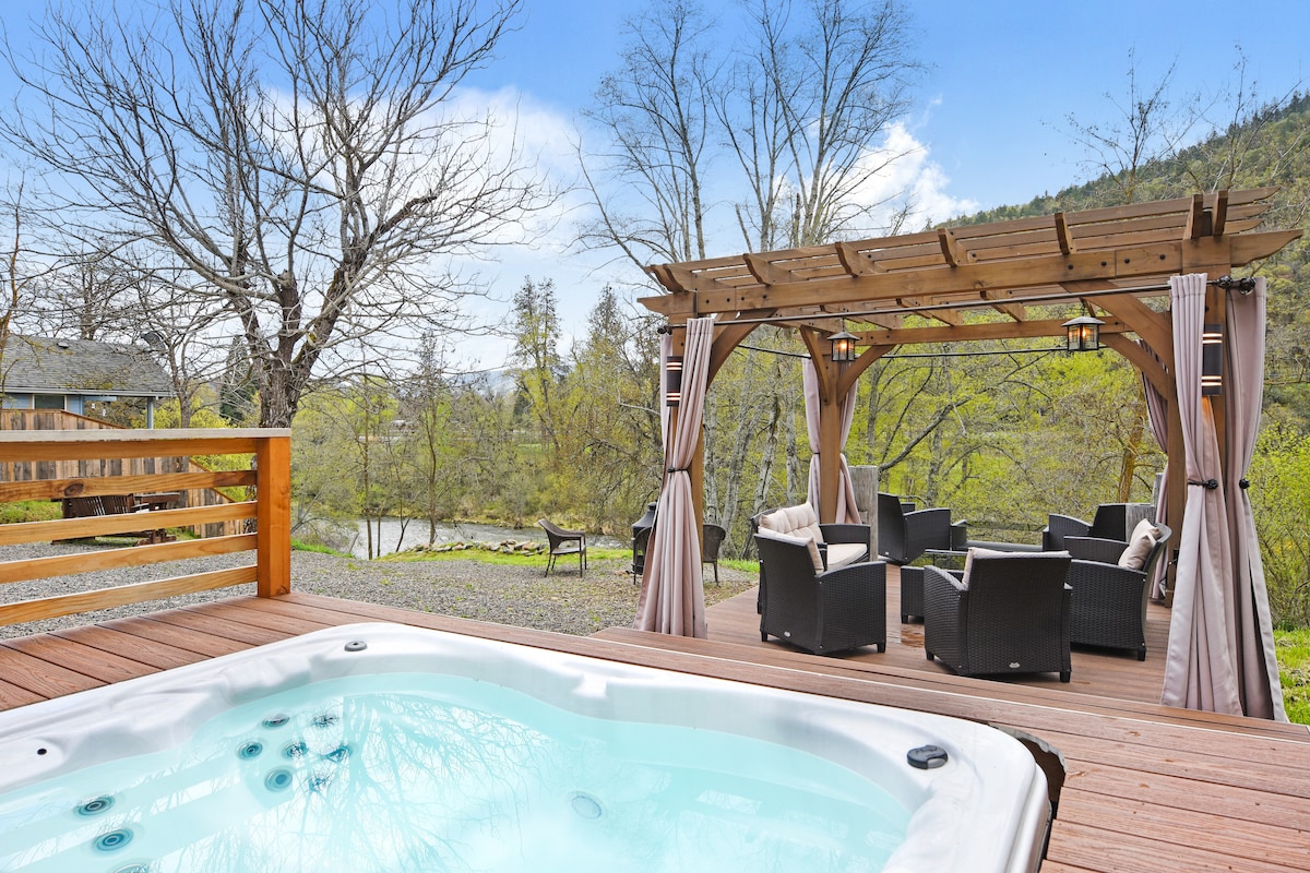 A hot tub is featured in the foreground, with bubbles gently rising. In the background, a covered seating area is visible, surrounded by trees. The landscape includes a glimpse of the river, framed by greenery and mountains.