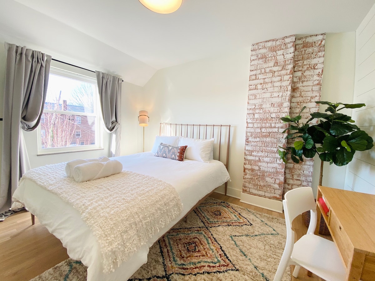 Segundo dormitorio en la planta superior con una extravagante cama tamaño queen, ladrillo blanco a la vista, cortinas oscuras, generoso armario con portaequipajes y perchas. Además, un televisor inteligente y un escritorio para un segundo espacio de trabajo.