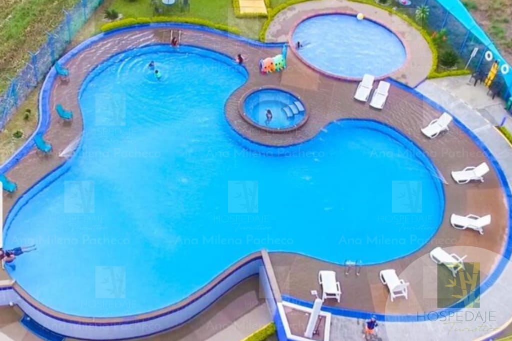 A spacious outdoor pool area features a unique shape with distinct sections for swimming, including a circular hot tub. Several lounge chairs surround the pool, inviting relaxation. The vibrant blue water is complemented by greenery and colorful play features for children, enhancing the recreational atmosphere.