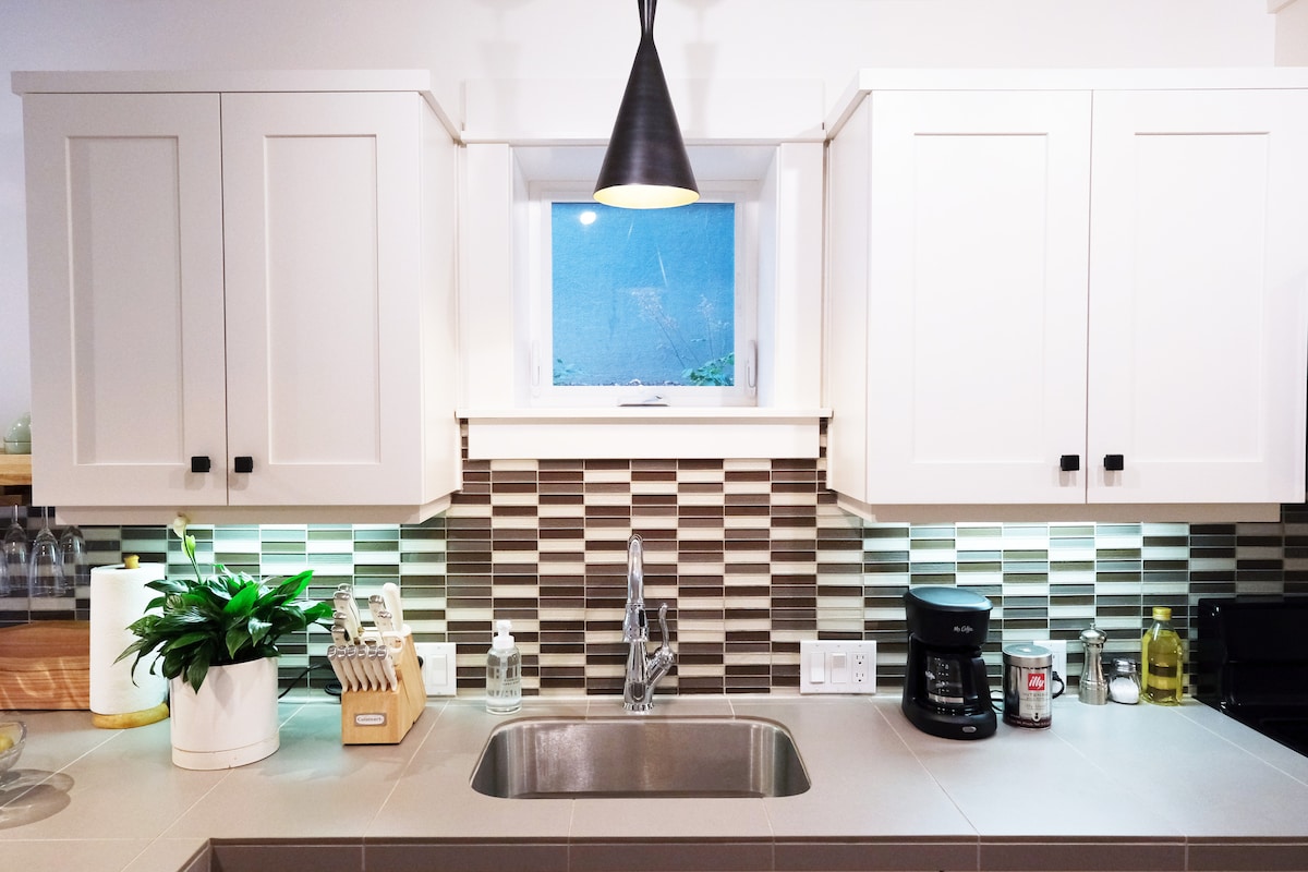 A modern kitchen features sleek cabinetry with overhead storage. Silver accents are found in the faucet and coffee maker, while a small plant adds a touch of greenery. A patterned backsplash and under-cabinet lighting enhance the inviting workspace, highlighting a functional kitchen area.