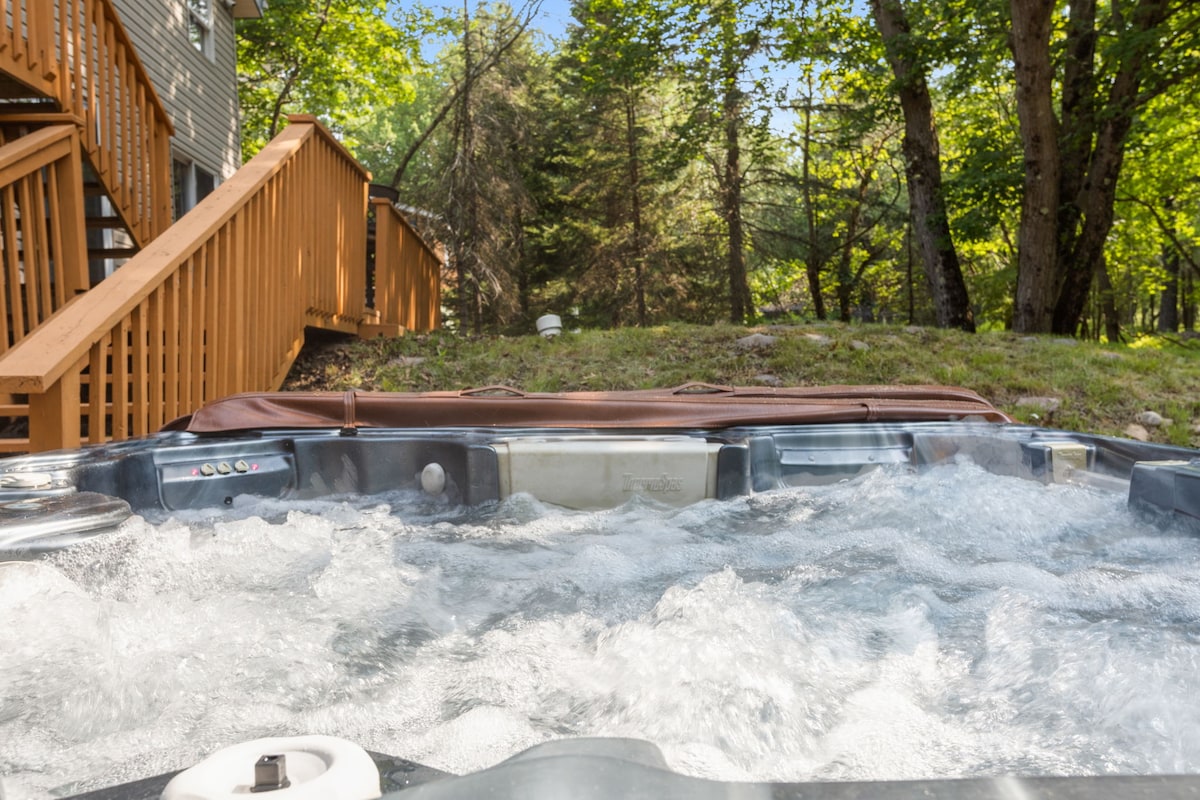 A five-person hot tub is featured, with bubbling water visible, set against a backdrop of lush greenery. Wooden steps leading up from the tub to the main building are included, enhancing the serene outdoor space.