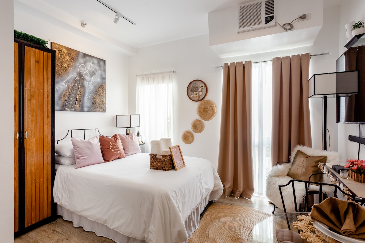 A cozy studio space features a spacious bed with decorative pillows in soft hues. Natural light filters through sheer and opaque curtains, illuminating the room. A wooden closet and a small desk with a chair add functionality, while wall decor includes woven pieces for visual interest.