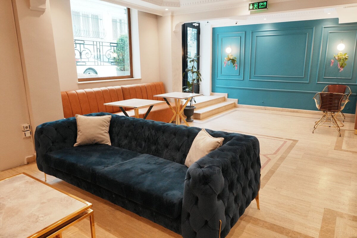 A spacious lobby is highlighted by a deep blue velvet sofa adorned with two neutral pillows. A glass-top table with gold accents complements the seating area, while green plants are visible near the window. Elegant wall decorations and a large staircase enhance the modern aesthetic.