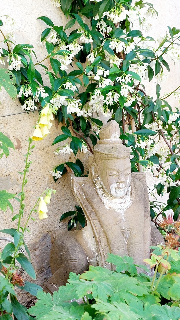 A serene garden setting features a partially obscured stone statue nestled against a textured wall. Lush green foliage and delicate white flowers surround the statue, creating a sense of tranquility and natural beauty.