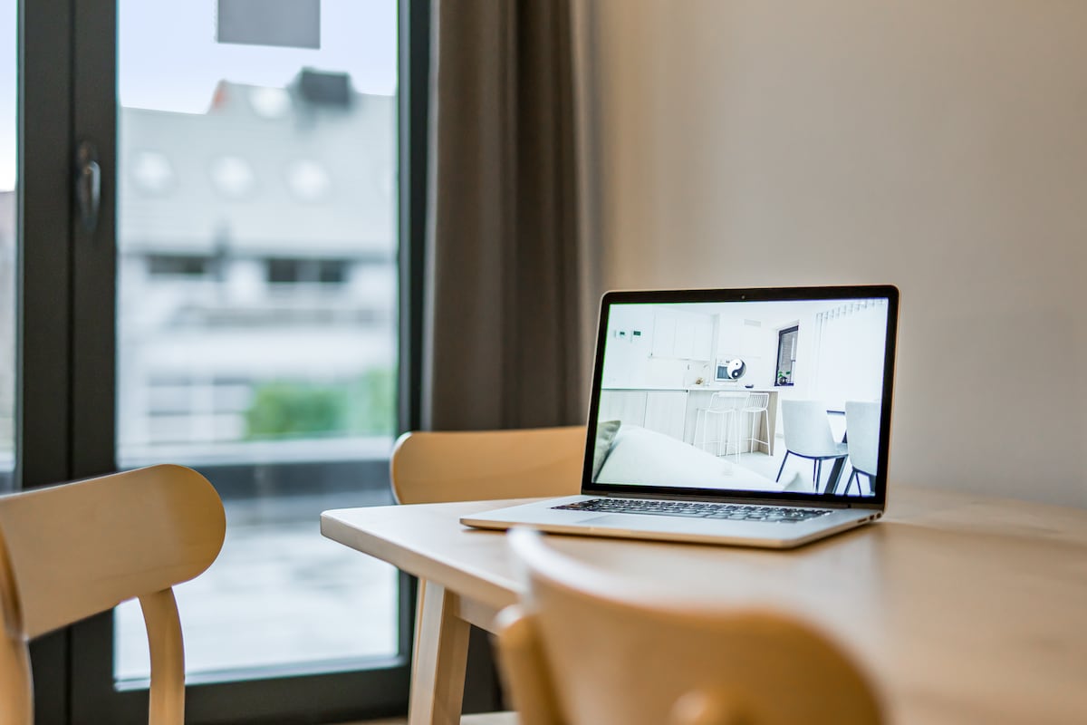 A clean workspace is seen on a wooden table, featuring a laptop open with a light-colored background. Two wooden chairs are positioned around the table, and a large window allows natural light to illuminate the area.