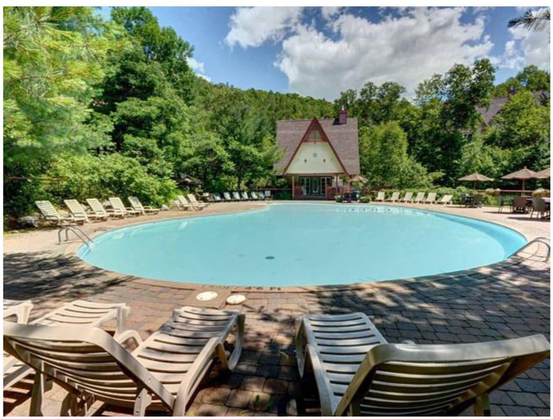 A spacious outdoor pool is surrounded by a landscaped area with tall trees. Sun loungers line one side of the pool, providing ample seating. The pool area is complemented by a charming building in the background, enhancing the inviting atmosphere of the complex.