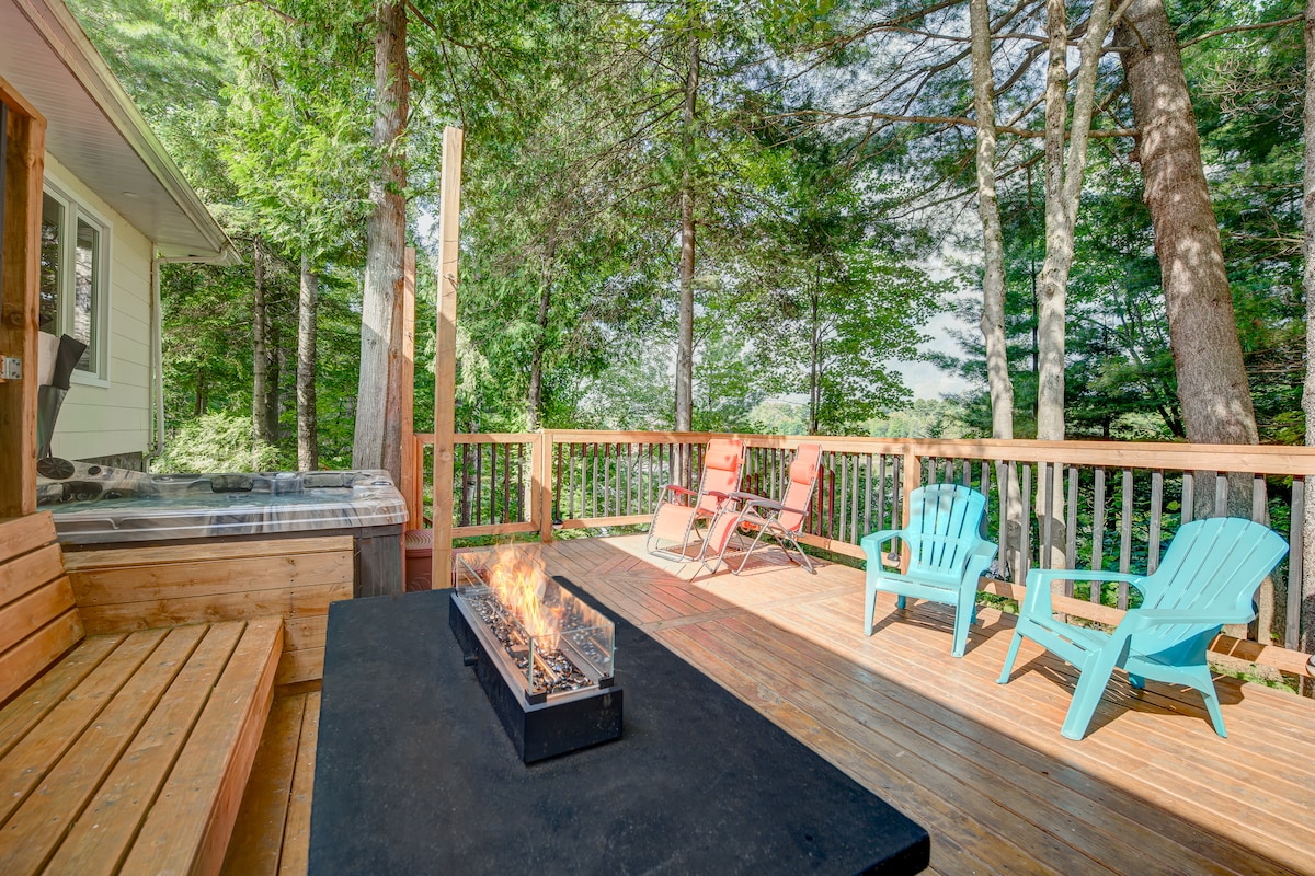 An outdoor terrace features wooden decking surrounded by tall trees, offering a secluded atmosphere. Seating arrangements include colorful chairs and a built-in bench. A rectangular fire pit sits at the center, with flames gently flickering, while a hot tub is visible at one end of the terrace.