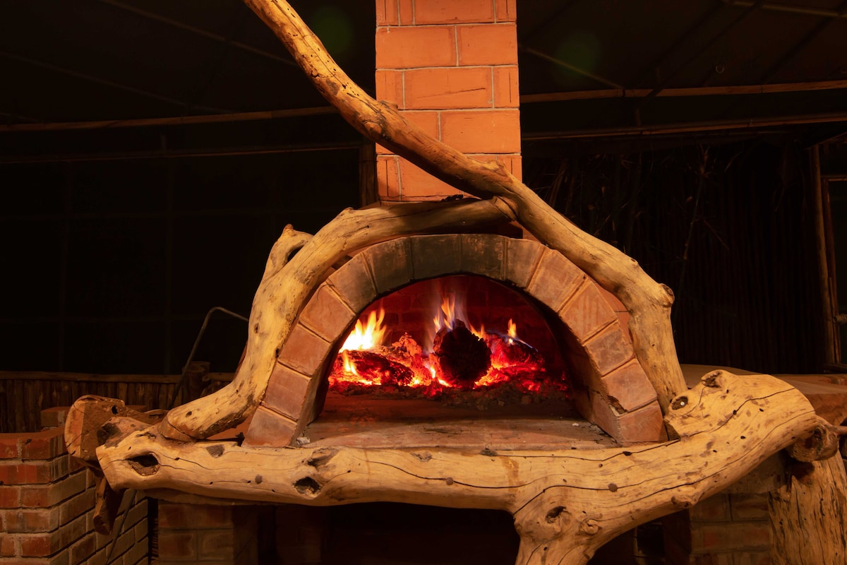 A rustic wood-fired oven is displayed, featuring a brick chimney and a curved entrance. Flames are visible inside, surrounded by glowing embers. The oven is framed by natural wooden elements, enhancing the camp's earthy aesthetic.