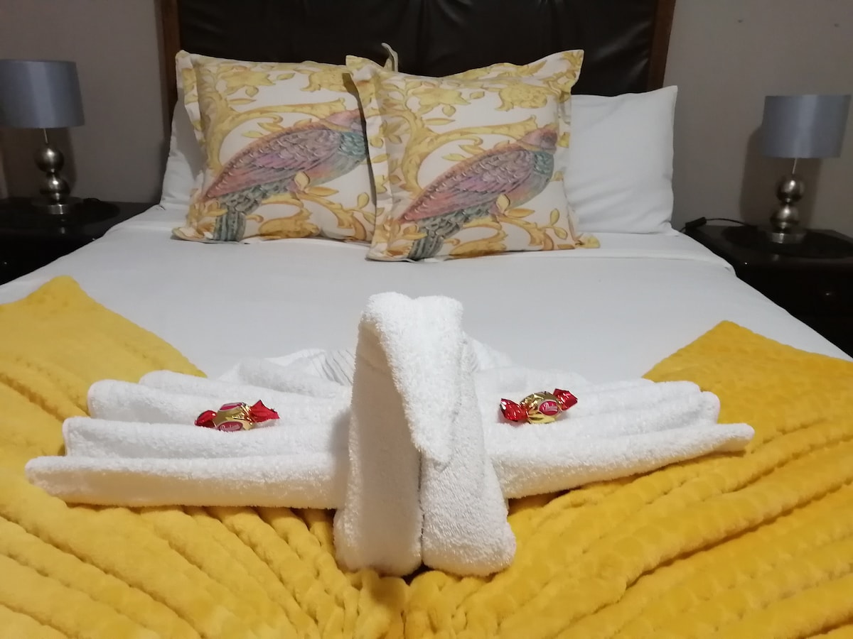A neatly arranged bed features decorative pillows with a bird motif and a cozy yellow throw blanket. Two towel sculptures are crafted into swans at the foot of the bed, complemented by chocolate treats placed nearby.