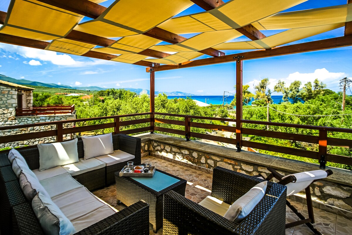 A shaded terrace features comfortable seating arranged around a coffee table. The warm tone of the woven furniture contrasts with the vibrant blue sea and greenery visible in the background, while a partially shaded overhead canopy allows for a relaxing outdoor space.
