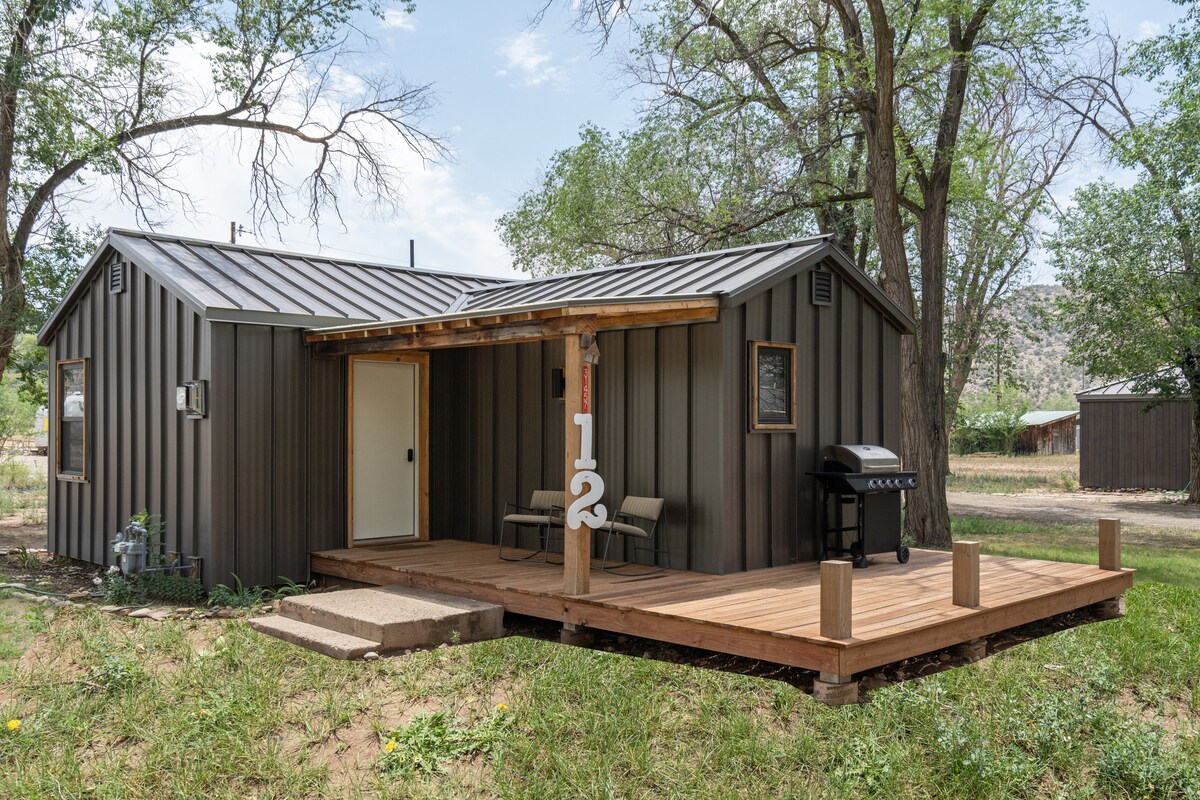 A rustic cabin is seen with dark metal siding and a sloped roof. The entrance features a wooden deck with two chairs and a gas grill. The number '12' is displayed prominently by the entrance, surrounded by greenery and trees.