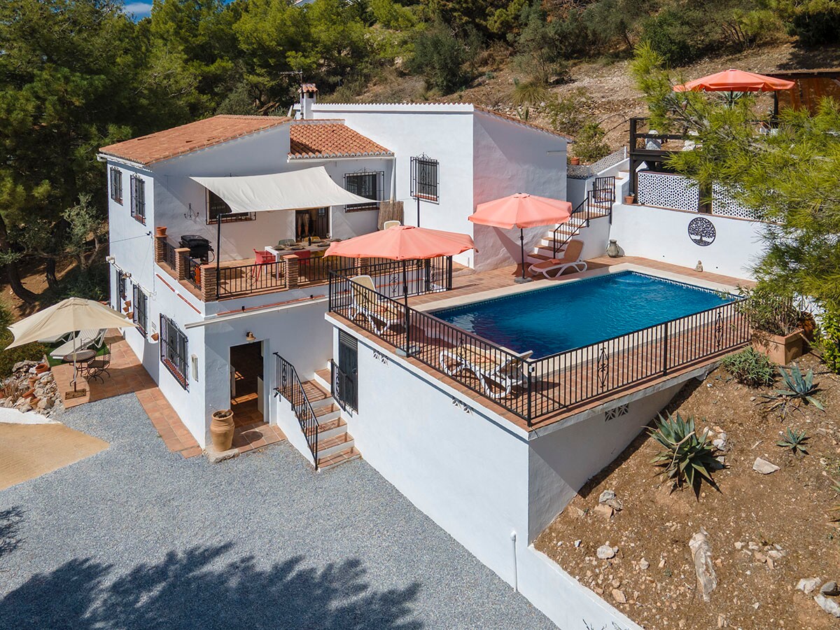 The villa is showcased from an elevated view, featuring a private pool surrounded by sun loungers and umbrellas. The spacious terraces provide areas for outdoor relaxation and dining, while the white exterior walls and tiled roof blend harmoniously into the rural landscape.