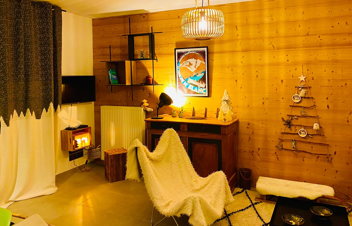 A cozy living area features wooden walls and a comfortable seating arrangement. A stylish light fixture hangs above a decorative cabinet, while a tree made of branches adds a rustic touch. A TV and bookshelves are also visible, creating an inviting space.