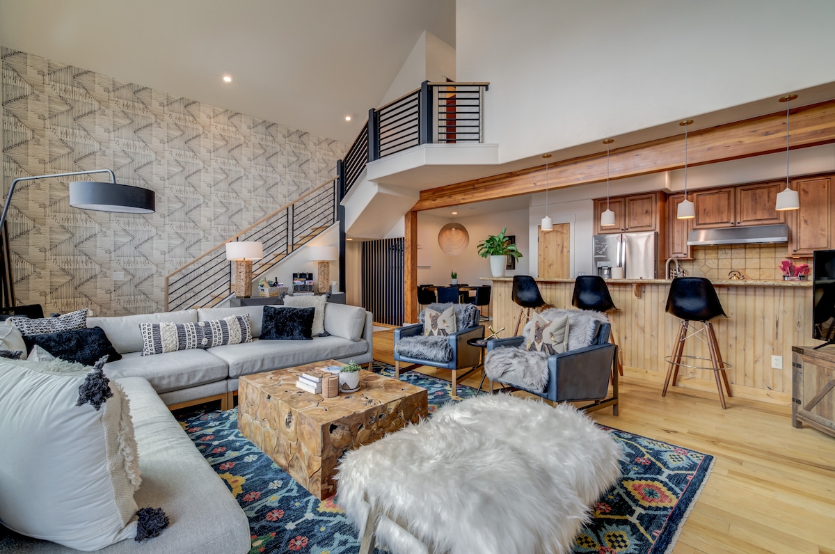 An open-concept great room is featured, showcasing hardwood floors and cathedral ceilings. A spacious sectional sofa, accented by decorative pillows, faces a unique wood coffee table. Bar stools line the kitchen island, and a warm ambiance is created by natural light flowing in from large windows.