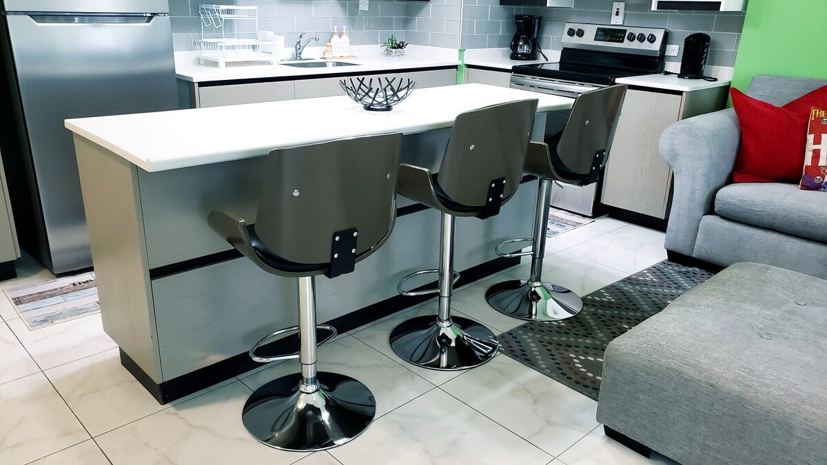 A sleek kitchen area is shown with modern bar stools positioned around a white countertop island. Stainless steel appliances and grey cabinetry complement the contemporary design. Light-colored tile flooring enhances the openness of the space, while a cozy couch is visible in the background.