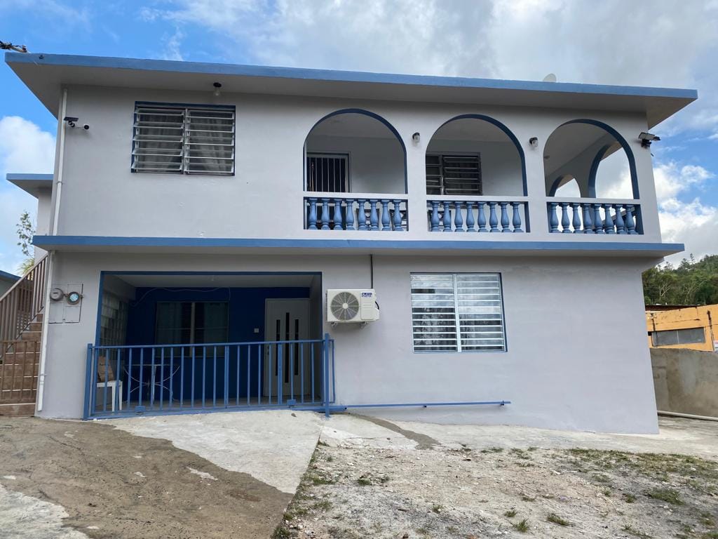 The exterior of a two-story building is presented, featuring a light-colored façade with blue accents. Arched balconies are visible on the upper level, complemented by white railings. A ground-level porch area is enclosed by a low blue fence, and a window air conditioning unit is mounted on one side.