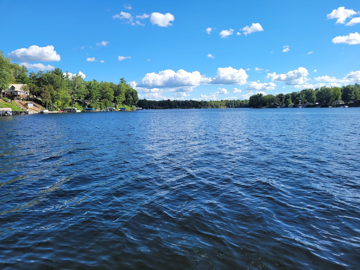 A serene view of Croton Pond showcases calm, rippling water under a bright blue sky scattered with fluffy clouds. Lush green trees line the shoreline, with hints of cottage structures visible in the distance, highlighting the natural beauty of the surroundings.