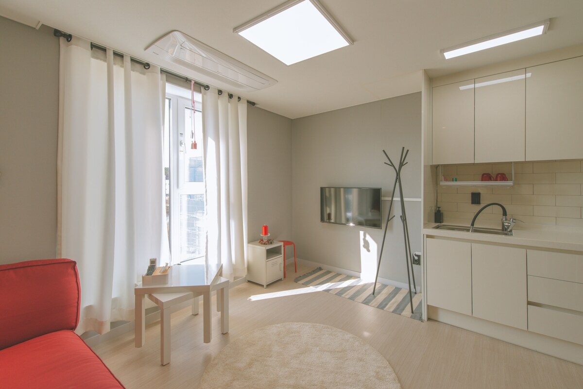 A bright living space with large windows allowing natural light to fill the room. A red couch is positioned next to a small coffee table, with a sleek television mounted on the wall. An open kitchen area features modern cabinetry and a minimalist design, enhancing the inviting atmosphere.