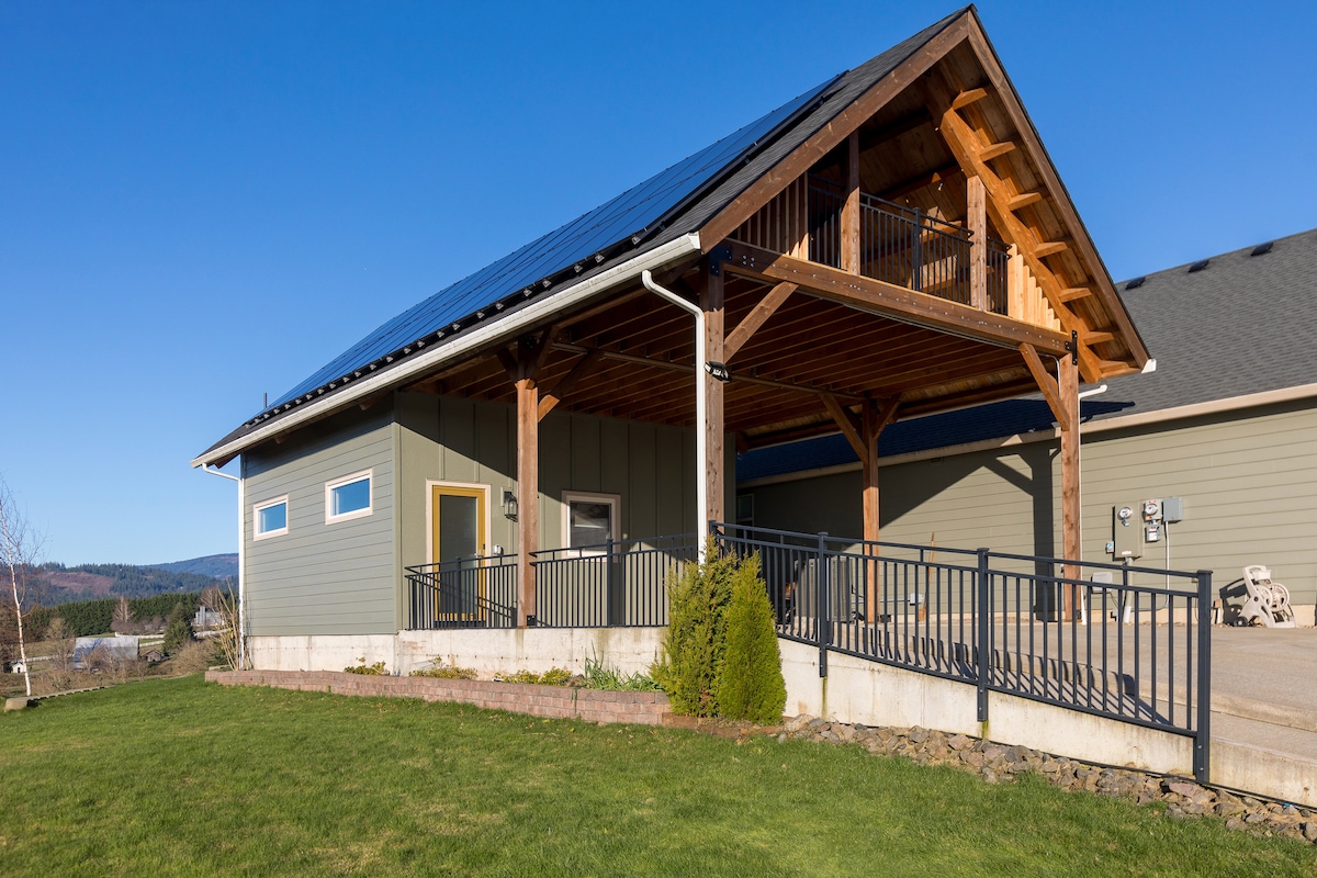 The exterior of a newly constructed cabin features a wooden deck supported by exposed beams. Solar panels are visible on the roof. A manicured lawn extends in front of the cabin, complemented by landscaped flower beds. Clear blue skies enhance the serene environment.