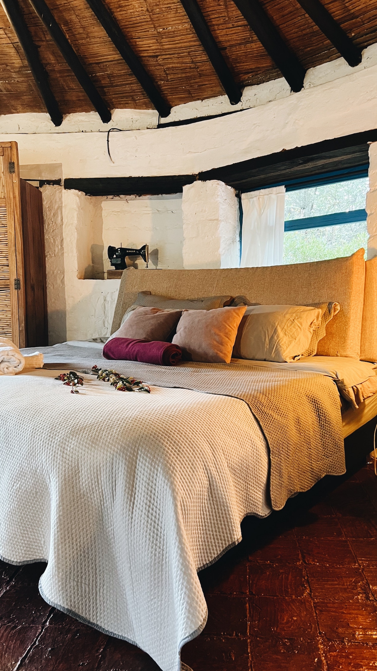 A cozy sleeping area features a spacious bed with neutral-toned bedding and decorative pillows. Natural light enters through a window draped in sheer curtains, illuminating the textured walls and wooden accents of the circular room.