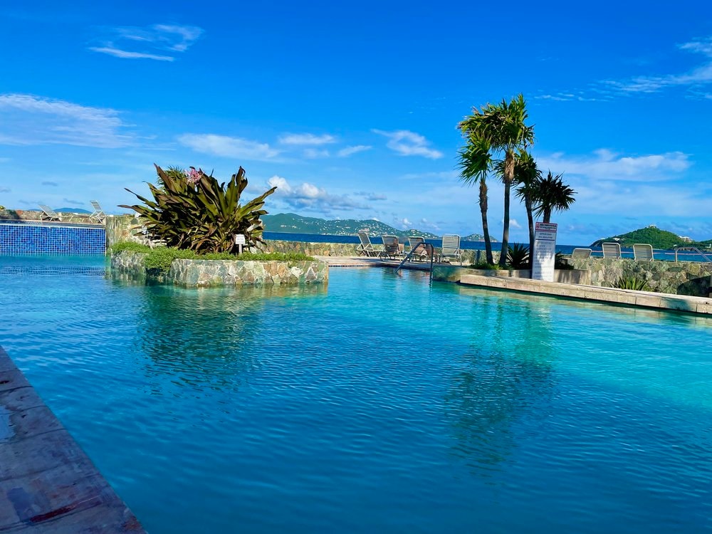A serene pool area is highlighted, surrounded by tropical plants and lounge chairs. The water reflects a vibrant blue sky, while palm trees provide a touch of greenery. The pool appears to extend towards the horizon, revealing a view of distant hills.